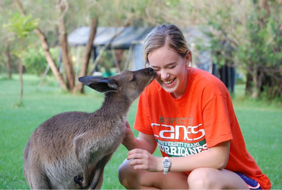 A student crouched beside a kangaroo laughs as the kangaroo sniffs her face.